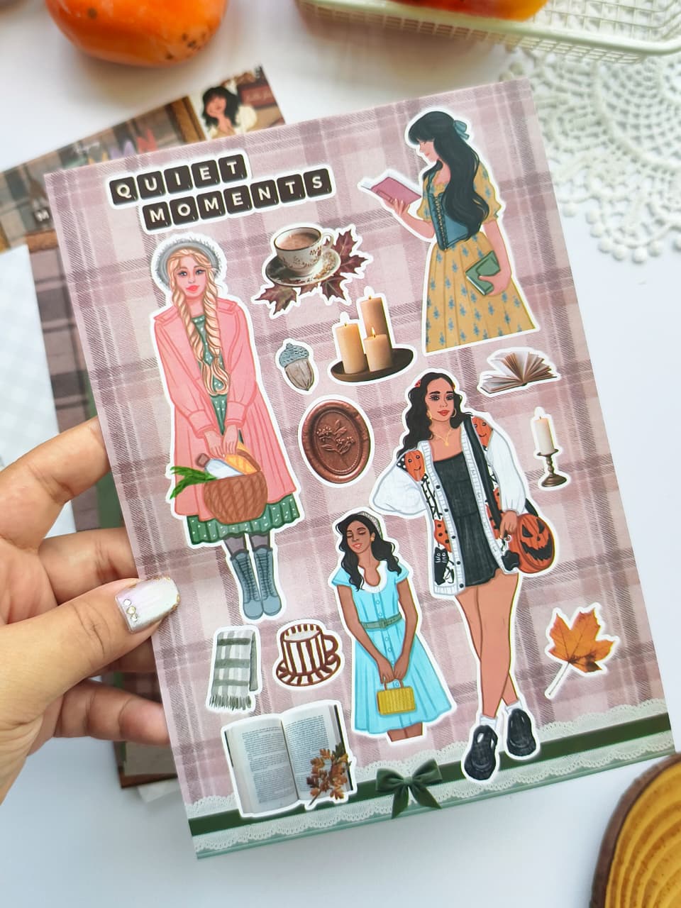 Hand holding the Autumn Moments vinyl sticker sheet featuring stylish autumn girls, books, candles, coffee, scarves, leaves, and cozy seasonal illustrations for planners, journals, and scrapbooks.