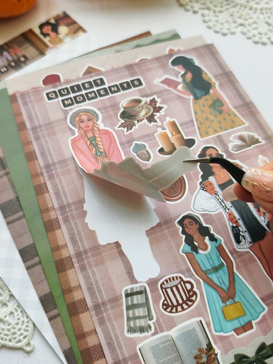 Close-up of the Autumn Moments vinyl sticker sheet showing a premium kiss-cut sticker being peeled away, featuring cozy autumn illustrations, book lovers, coffee, candles, and fall-inspired accessories.