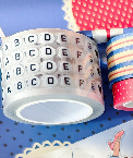 A close-up of a roll of decorative tape with printed letters A through F on a white background, alongside other rolls of patterned tape in red, blue, and white.