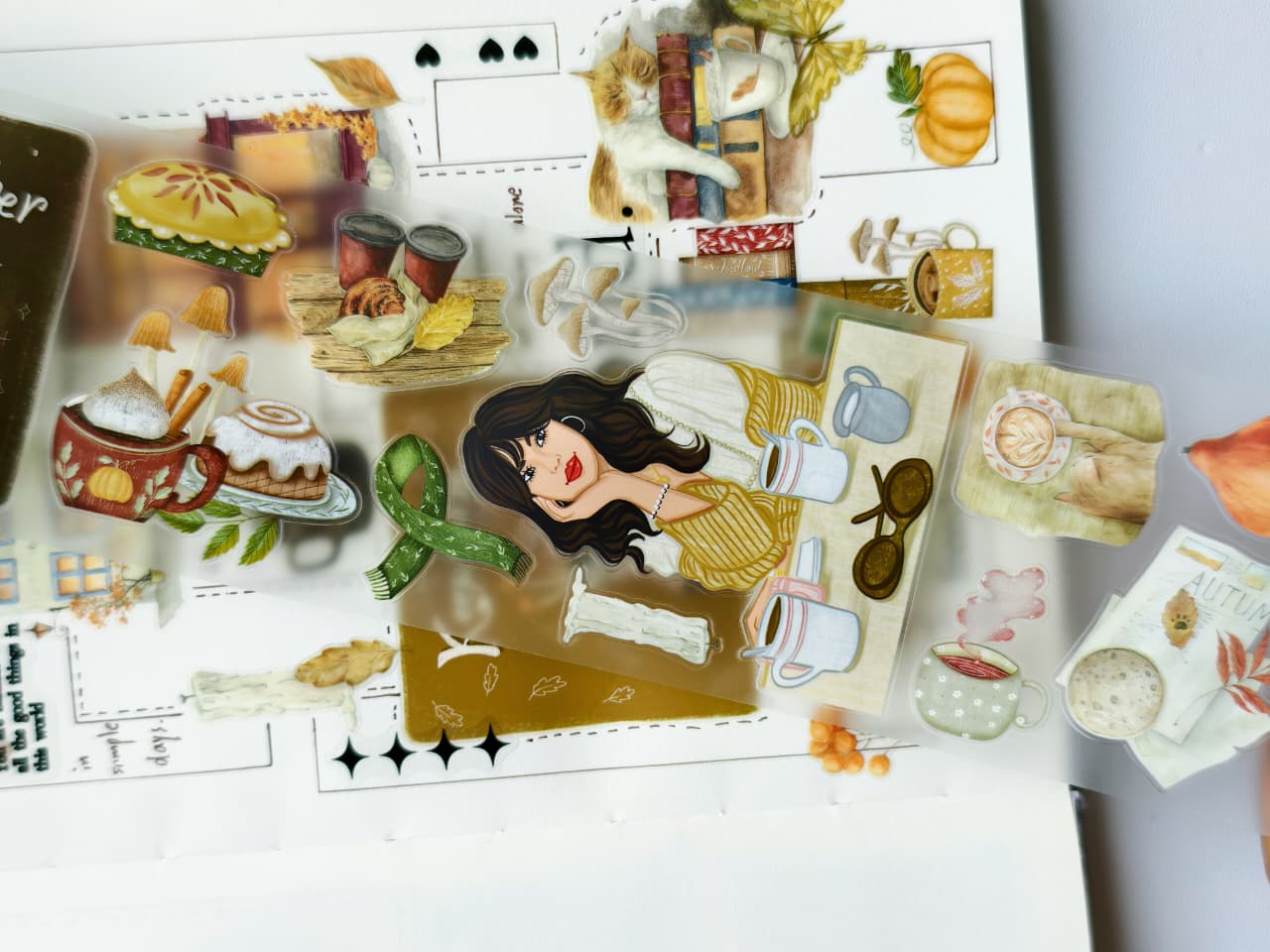 Close-up of transparent pre-cut PET stickers featuring coffee mugs, pastries, books, pumpkins, cozy clothing, autumn décor, and a stylish woman enjoying a relaxing fall day.