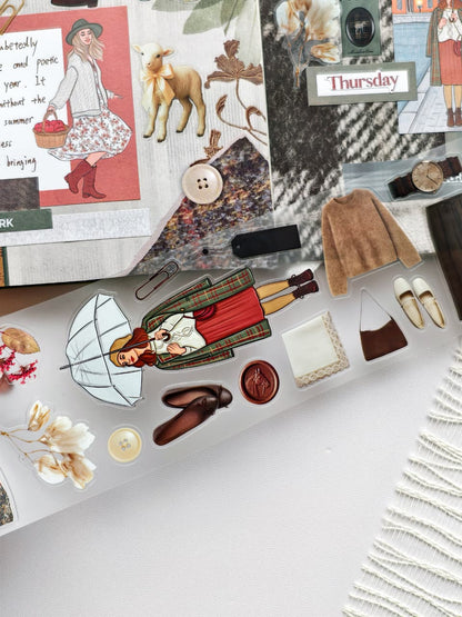 Close-up of Autumn Tales PET tape showcasing a red-haired girl holding a clear umbrella, cozy knit sweater, vintage shoes, plaid coat, lamb illustration, buttons, wax seals, and autumn-themed decorative elements.