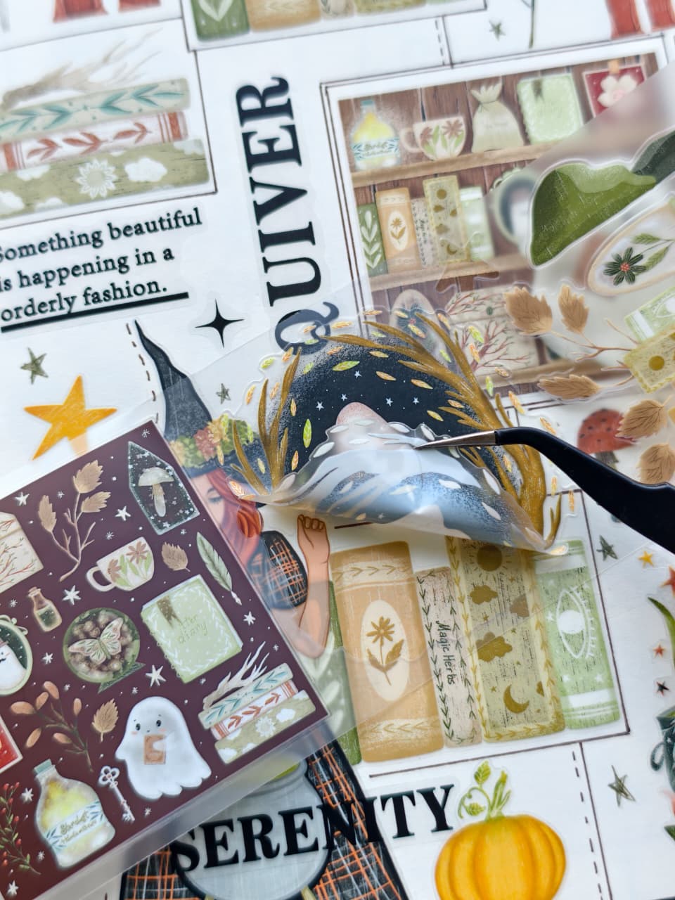 Close-up of transparent pre-cut stickers featuring a ghost floating inside a golden wreath, magical books, autumn foliage, potion bottles, and whimsical Halloween-inspired artwork being lifted with tweezers.