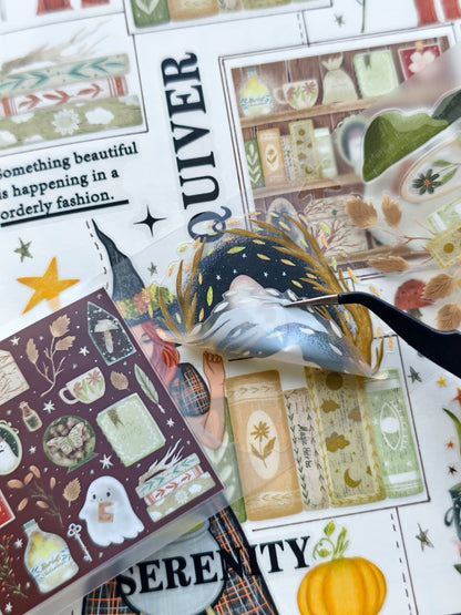 Close-up of transparent pre-cut stickers featuring a ghost floating inside a golden wreath, magical books, autumn foliage, potion bottles, and whimsical Halloween-inspired artwork being lifted with tweezers.