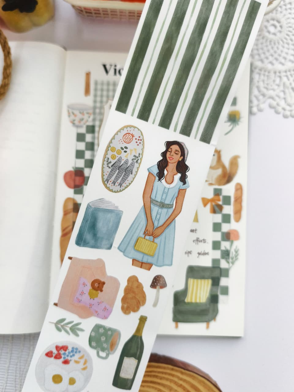 Close-up of Cozy Cottage Washi Tape featuring watercolor illustrations of a woman in a blue dress, breakfast, mushrooms, sweater, mug, bottle, croissant, striped washi pattern, and cozy cottage-inspired accents.