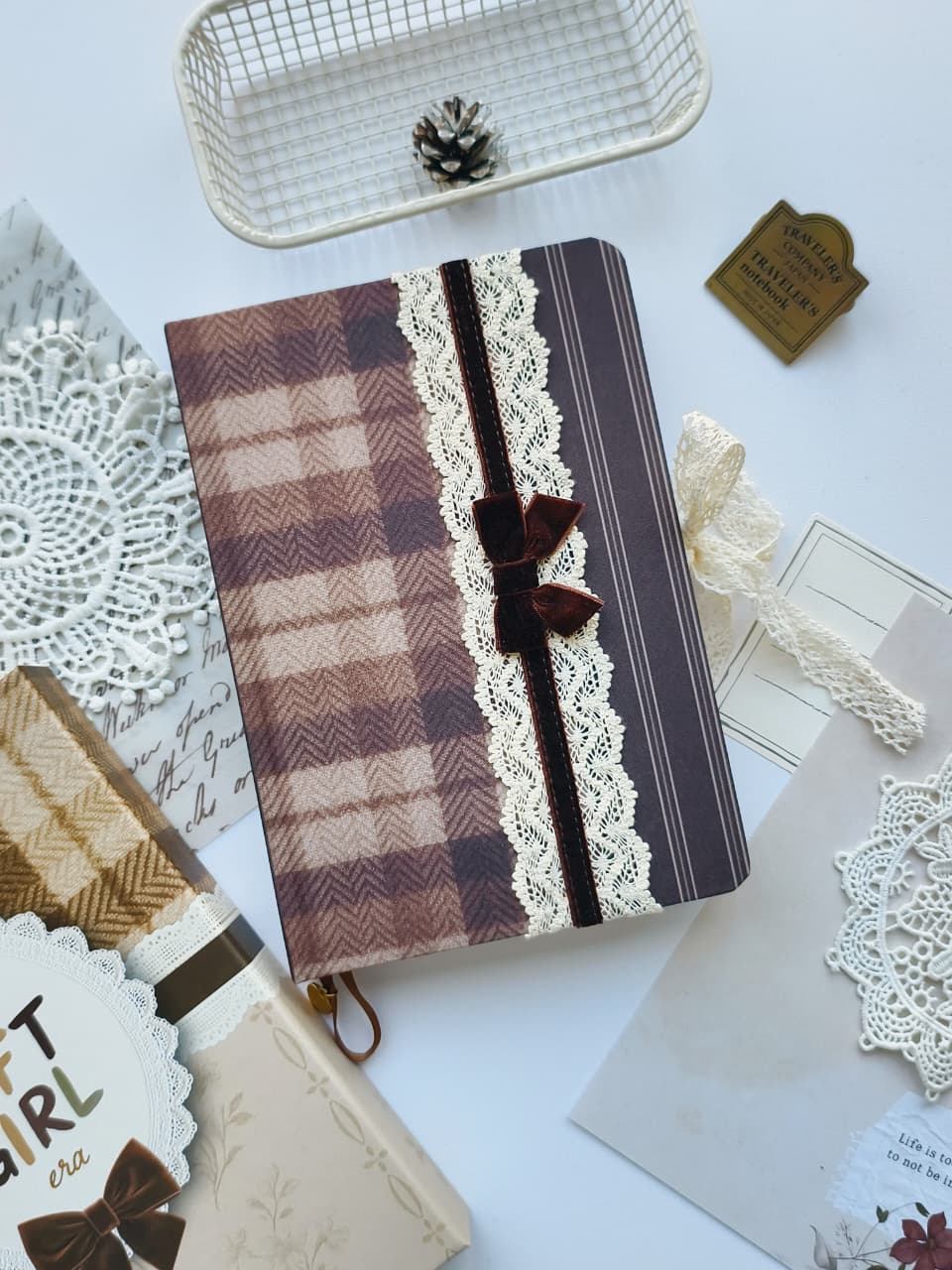 A decorative notebook with a brown and beige plaid cover, cream lace trim, and a dark brown ribbon bow, styled on a white surface with paper doilies, a small basket, and stationery props.