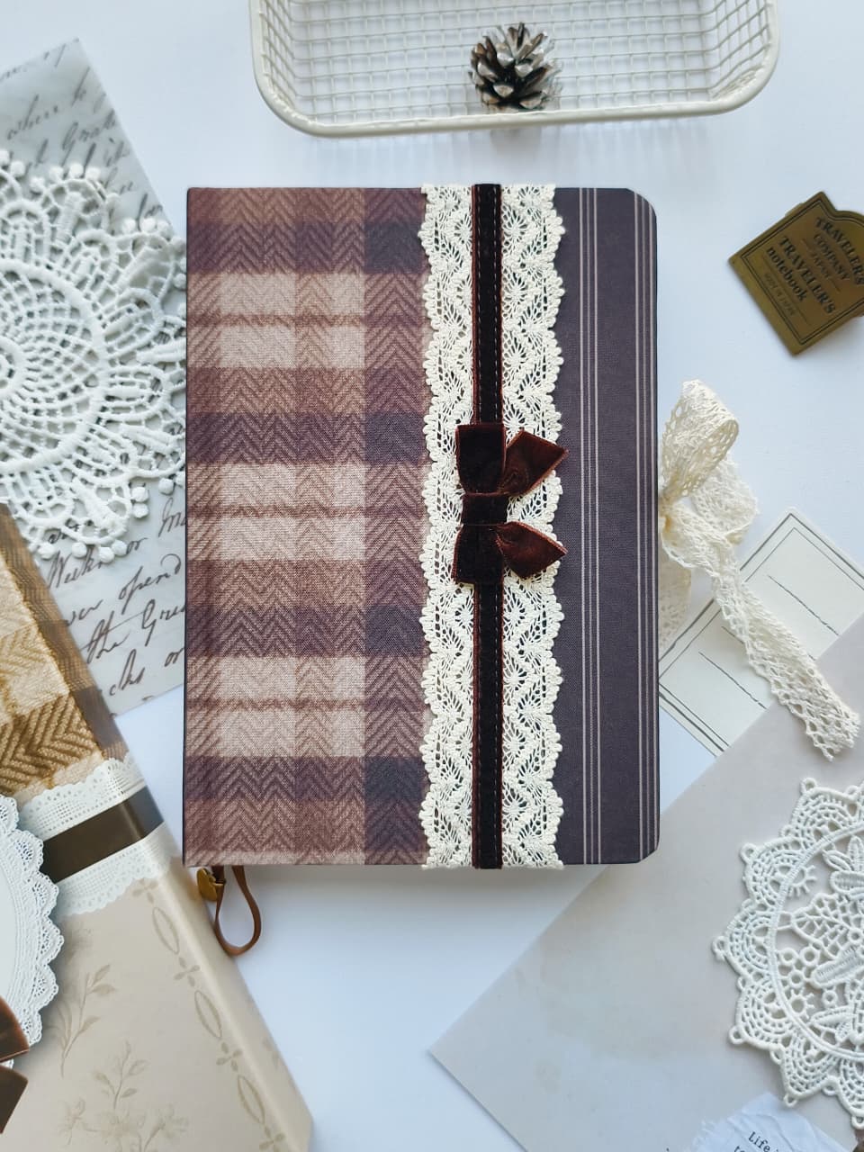 A decorative notebook or journal cover laid flat on a white surface, featuring a brown and beige plaid pattern, cream lace trim, a dark brown ribbon closure with a bow, and striped accents. Surrounding props include lace doilies, envelopes, a small pinecone in a tray, and a paper tag.