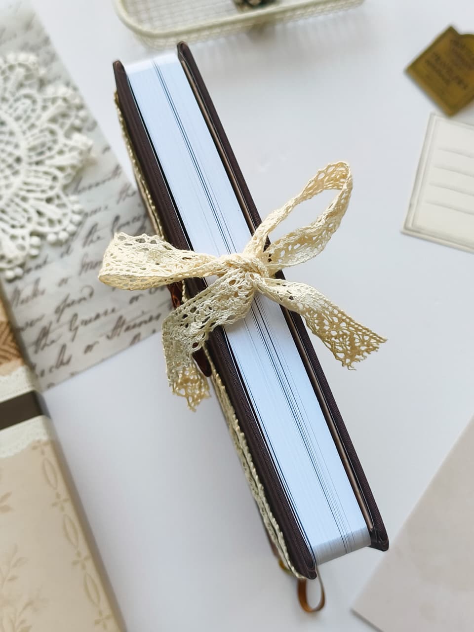 A vintage-style notebook with a dark brown leather cover and light blue pages, tied with a cream lace ribbon, displayed on a white surface with decorative stationery.