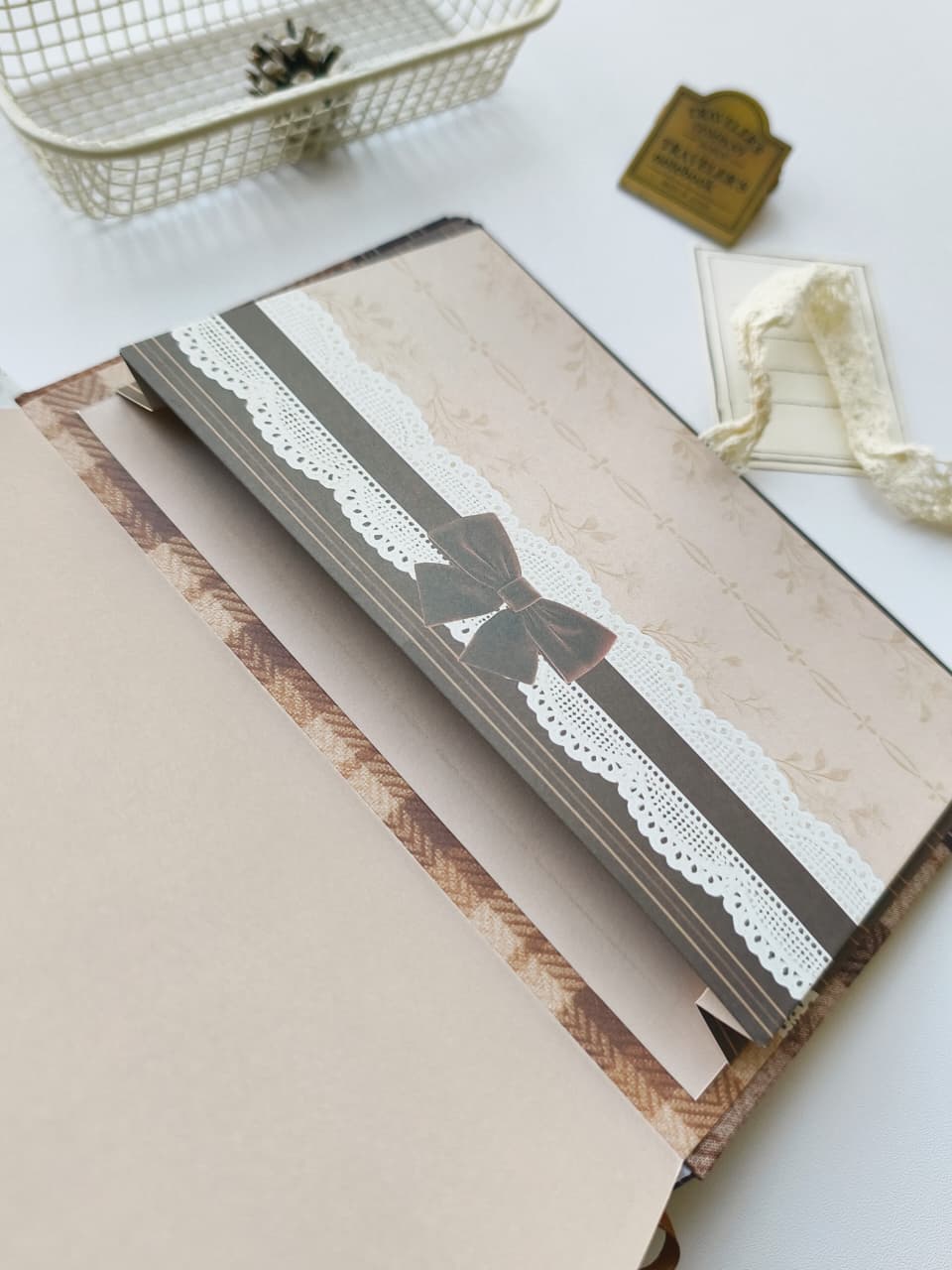 A vintage-style decorative notebook or journal cover laid open on a white surface, featuring a beige cover with a brown ribbon, white lace trim, and a bow detail. A matching beige page is visible inside, with small styled props including a wire basket, a small card, and ribbon in the background.