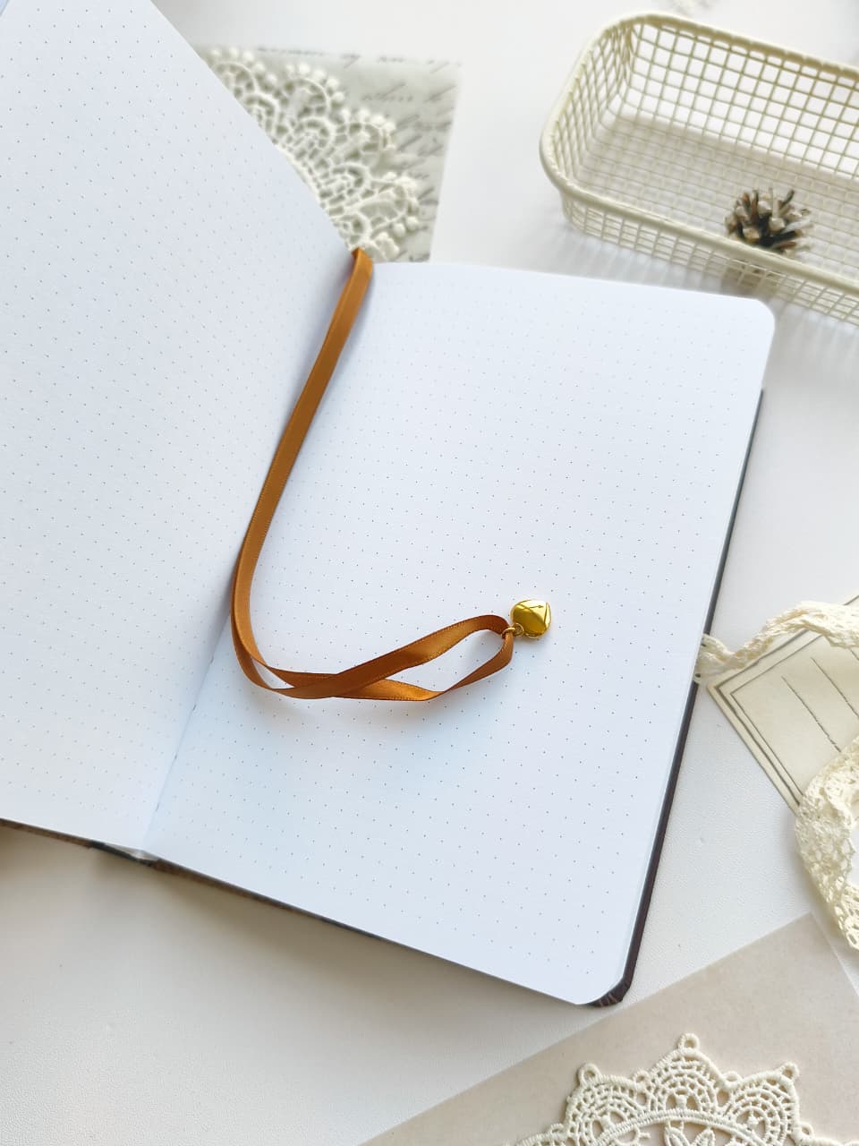 An open white dotted journal or notebook with a long tan ribbon bookmark draped across the pages and a small gold bead accent, styled on a light surface with lace and decorative props around it.