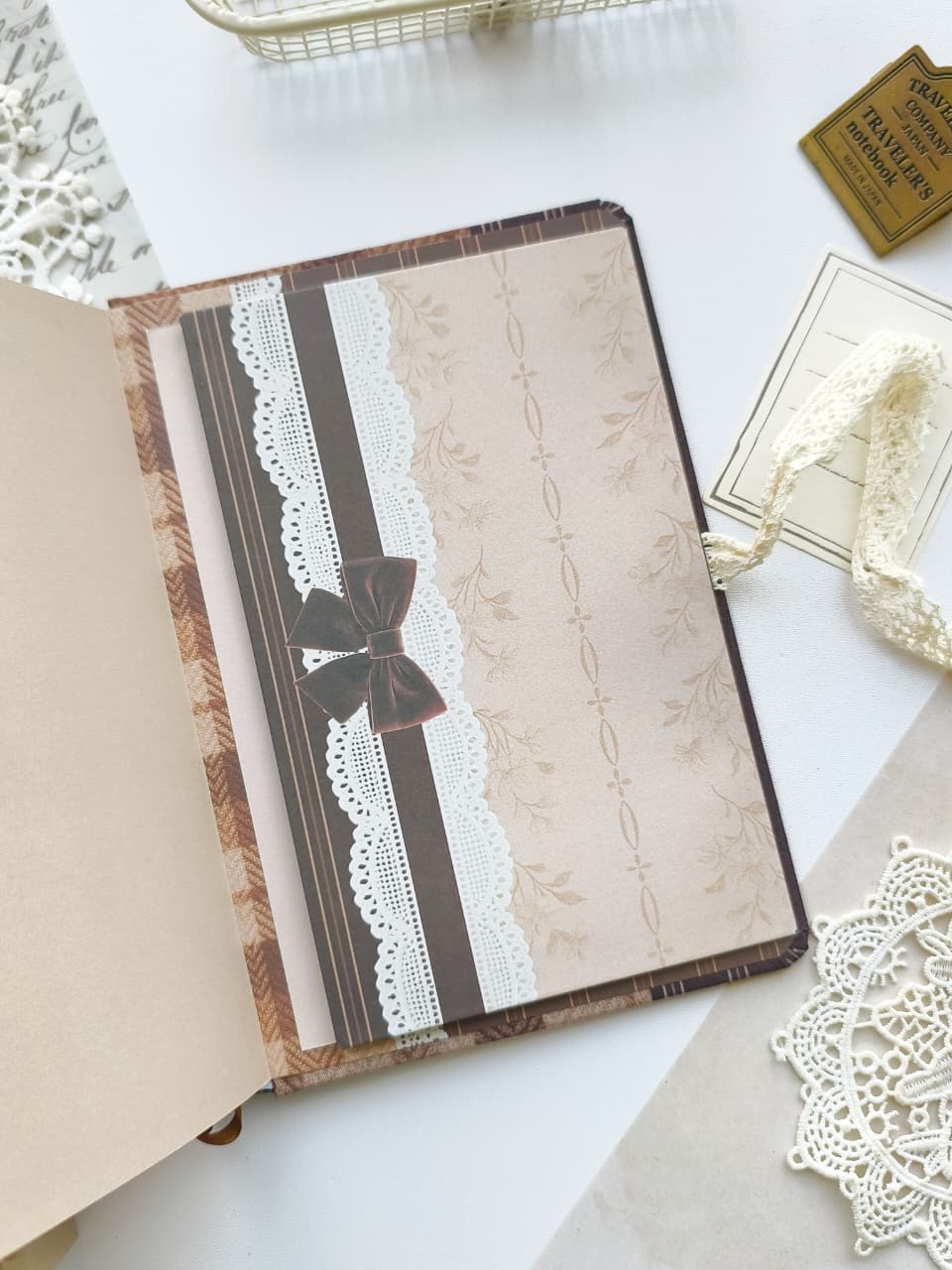 A vintage-style decorative notebook or journal shown open on a white surface, with a beige patterned cover, brown and white lace trim, and a dark brown bow closure. Surrounding props include lace, a small gold ticket, and paper elements.