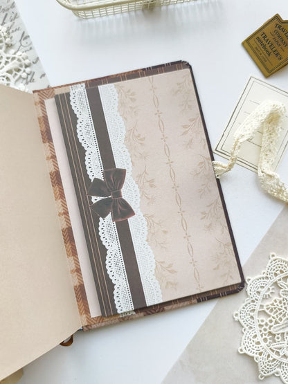 A vintage-style decorative notebook or journal shown open on a white surface, with a beige patterned cover, brown and white lace trim, and a dark brown bow closure. Surrounding props include lace, a small gold ticket, and paper elements.