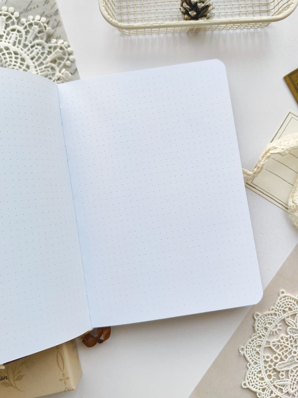 An open bullet journal with light blue dotted grid pages, styled on a white surface with lace doilies, a small pinecone in a wire basket, and neutral decorative props.