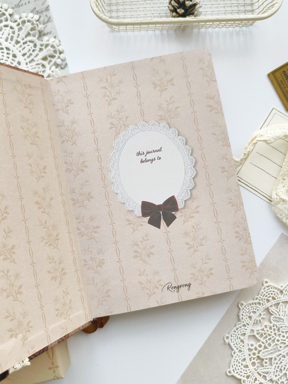 An open vintage-style keepsake journal with a beige floral patterned cover, featuring a central white oval insert with a lace border and a dark brown bow, surrounded by decorative lace and paper props on a white surface.