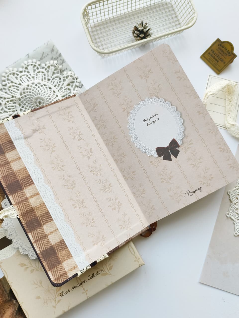 An open vintage-style notebook or journal on a white surface, showing patterned cream and beige pages with a circular white label that reads &