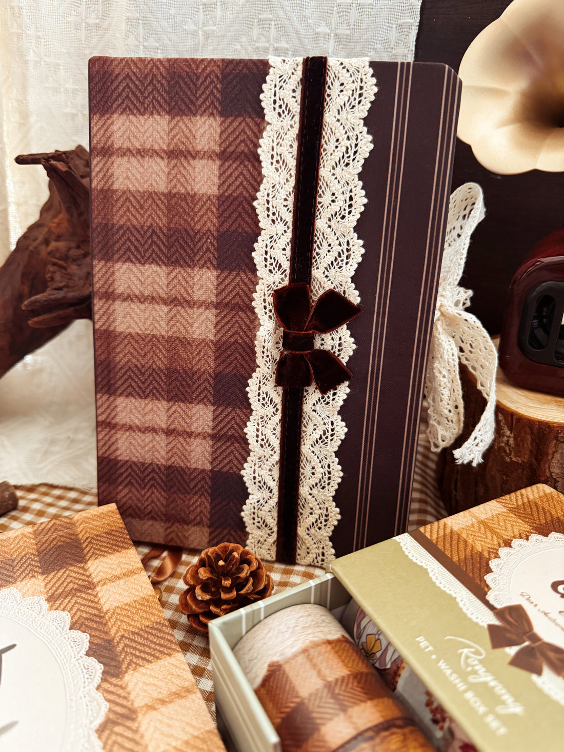 A styled product display featuring a brown and beige plaid notebook with a vertical white lace panel and a dark brown ribbon bow. The notebook is surrounded by matching autumn-themed packaging, a pinecone, and rustic props on a checkered fabric surface.