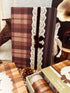 A styled product display featuring a brown and beige plaid notebook with a vertical white lace panel and a dark brown ribbon bow. The notebook is surrounded by matching autumn-themed packaging, a pinecone, and rustic props on a checkered fabric surface.