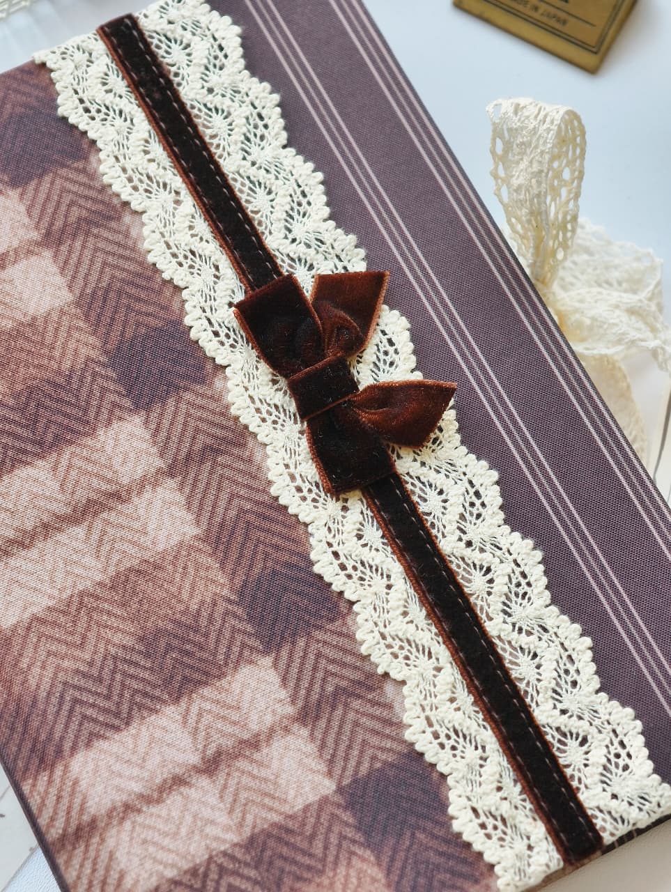 A close-up of a decorative fabric-covered item, likely a notebook, journal, or gift box, featuring a brown and beige herringbone pattern, a cream lace border, and a dark brown velvet ribbon bow with matching ribbon tails.