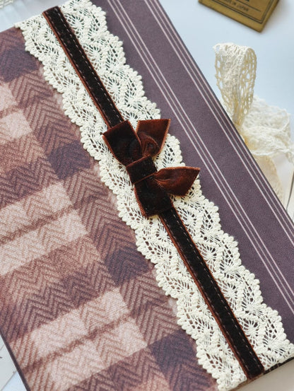 A close-up of a decorative fabric-covered item, likely a notebook, journal, or gift box, featuring a brown and beige herringbone pattern, a cream lace border, and a dark brown velvet ribbon bow with matching ribbon tails.
