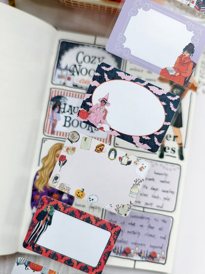 Close-up of Halloween-themed sticky notes showcasing ornate frames, spooky icons, and illustrated characters, displayed over a decorated journal spread with pumpkins and gothic accents.