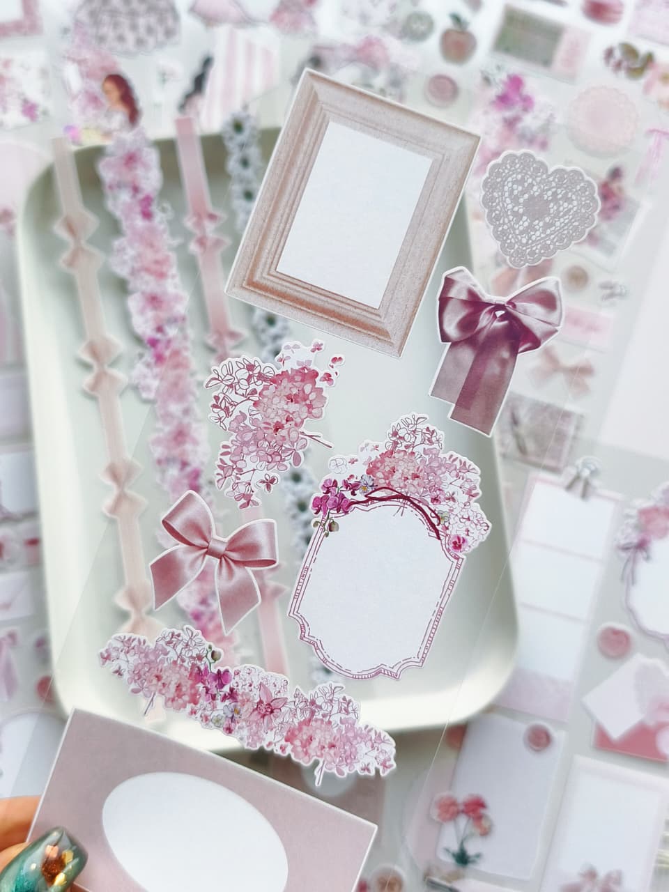 Close-up of transparent sticker sheet with pink floral clusters, satin bows, vintage frames, and delicate romantic journaling elements for planners and scrapbooks.
