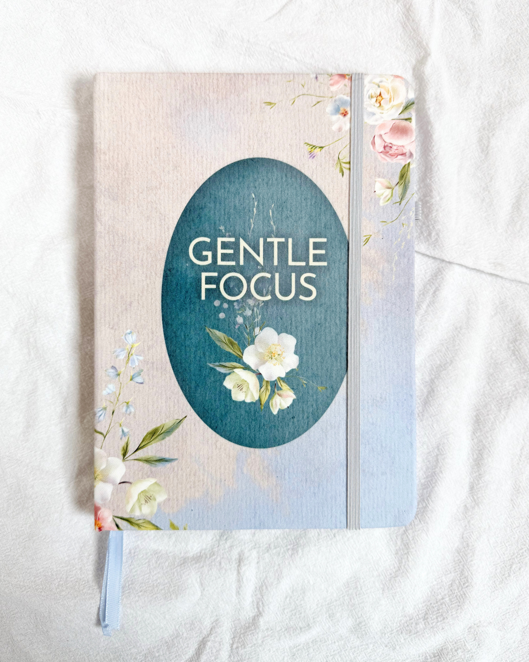 A floral hardcover journal titled "Gentle Focus" with a soft beige linen-style cover, featuring a teal oval title panel, delicate white and pink flowers, and a light gray elastic closure, photographed on a white fabric background.