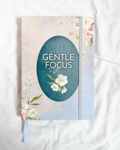 A floral hardcover journal titled "Gentle Focus" with a soft beige linen-style cover, featuring a teal oval title panel, delicate white and pink flowers, and a light gray elastic closure, photographed on a white fabric background.