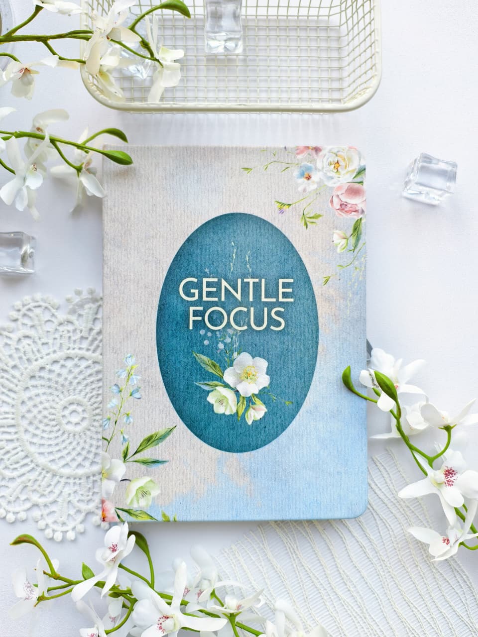 A floral Gentle Focus journal with a soft linen-textured cover, featuring a teal oval title panel and delicate white and pink flowers. The journal is styled on a bright white surface surrounded by white blossoms, lace, a glass tray, and decorative glass cubes, creating a clean, elegant, and serene flat-lay scene.