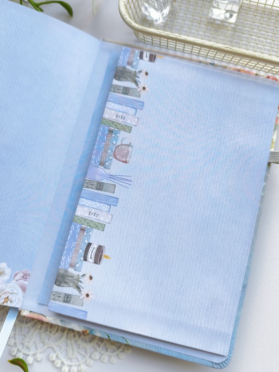 A close-up of the inside front cover of the Gentle Focus journal, featuring a soft blue interior with an illustrated bookshelf border along the edge. The journal is styled on a bright white surface with delicate floral accents, highlighting its premium paper and elegant design.
