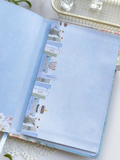 A close-up of the inside front cover of the Gentle Focus journal, featuring a soft blue interior with an illustrated bookshelf border along the edge. The journal is styled on a bright white surface with delicate floral accents, highlighting its premium paper and elegant design.