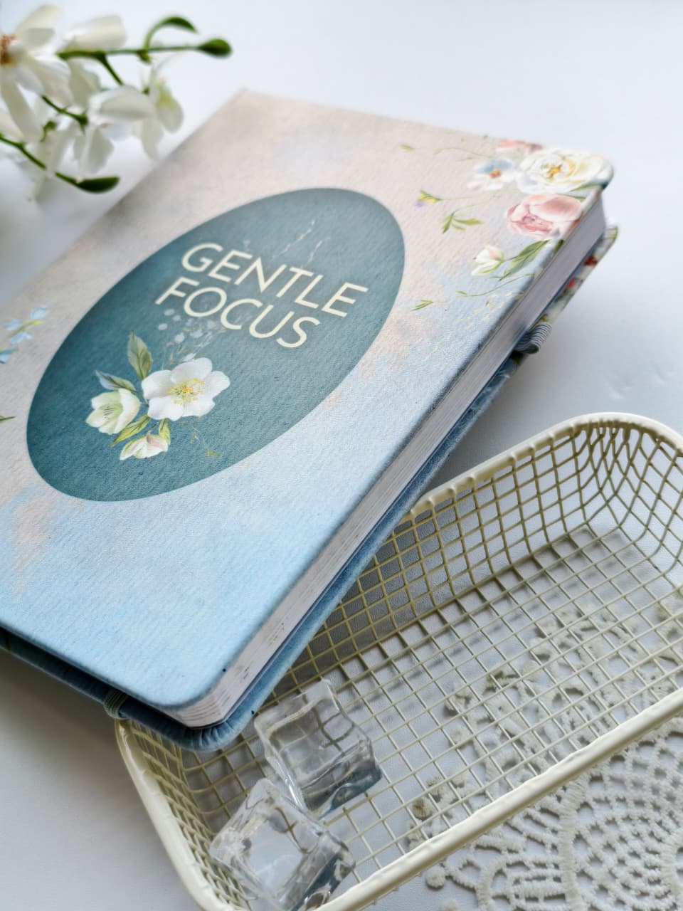 A close-up of the closed Gentle Focus journal showcasing its soft linen-textured floral cover with a teal title panel and delicate white and pink flowers. The journal is styled on a bright white surface with a decorative wire tray, lace accents, glass cubes, and white blossoms, highlighting its elegant design and premium hardcover finish.