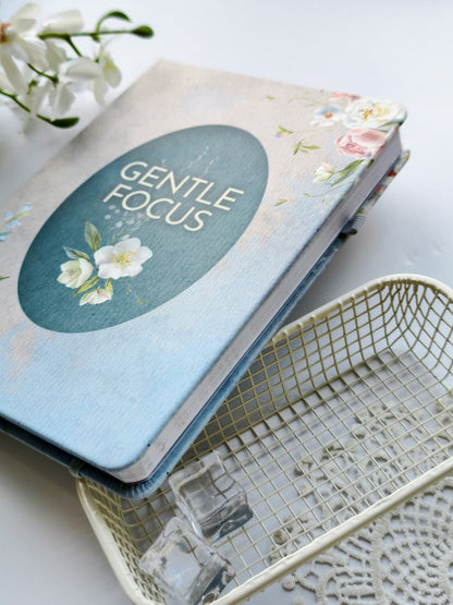 A close-up of the closed Gentle Focus journal showcasing its soft linen-textured floral cover with a teal title panel and delicate white and pink flowers. The journal is styled on a bright white surface with a decorative wire tray, lace accents, glass cubes, and white blossoms, highlighting its elegant design and premium hardcover finish.