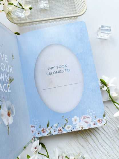 An open Gentle Focus journal displaying the personalized "This Book Belongs To" page, featuring a soft blue background, delicate floral illustrations, and an oval space to write the owner&
