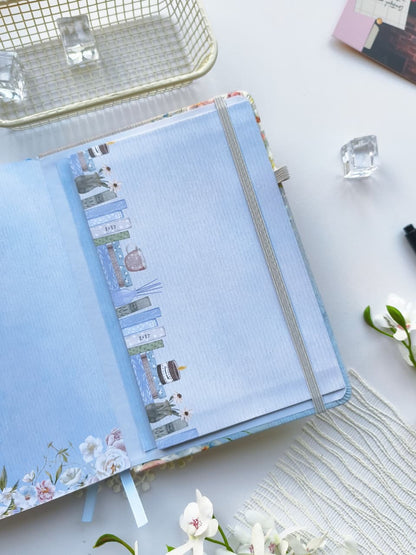 A close-up of the inside back cover of the Gentle Focus journal, featuring a built-in storage pocket with a charming illustrated bookshelf design and a secure gray elastic band. The journal is styled on a bright white surface with delicate floral accents, highlighting its elegant details and practical storage for notes and keepsakes.