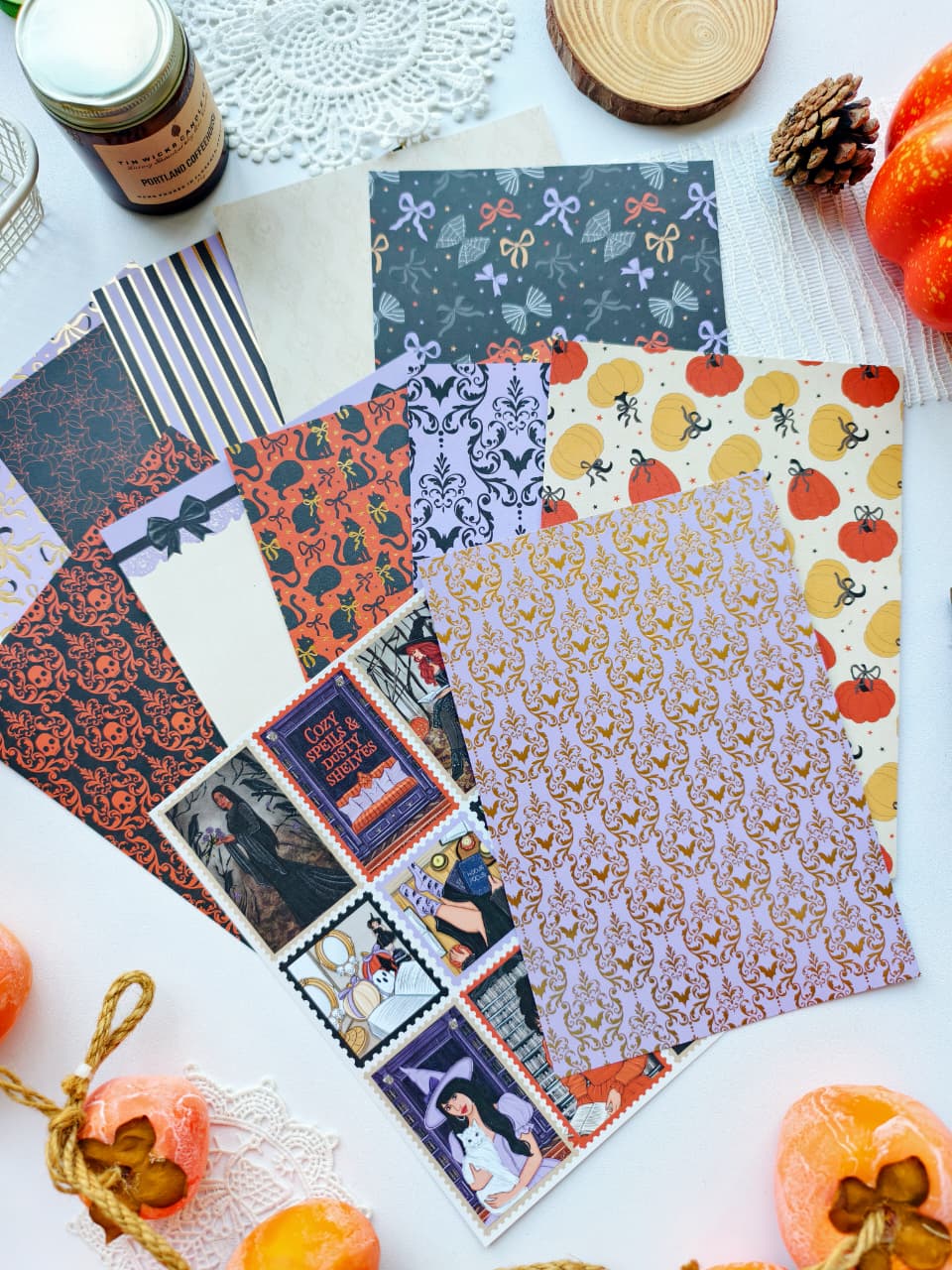 A styled flat-lay of multiple patterned paper sheets and a few printed cards arranged on a white surface with seasonal props including pumpkins, a pinecone, lace, and a small jar.