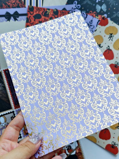 A hand holds a square sheet of lavender paper with a metallic gold damask pattern, with other patterned sheets visible in the background.