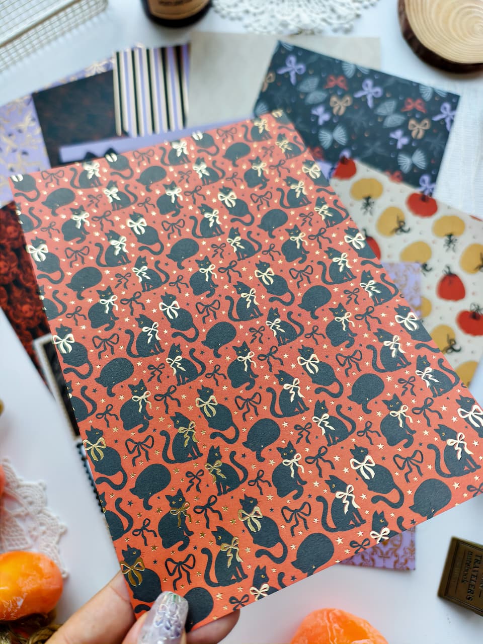 A hand holds a sheet of patterned craft paper featuring black cats wearing gold bows on an orange background, surrounded by other patterned papers and autumn-themed props.