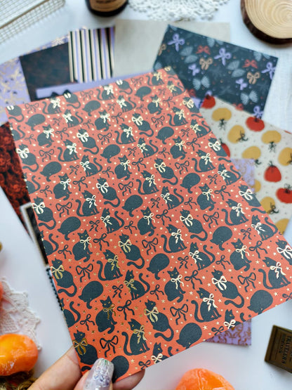 A hand holds a sheet of patterned craft paper featuring black cats wearing gold bows on an orange background, surrounded by other patterned papers and autumn-themed props.