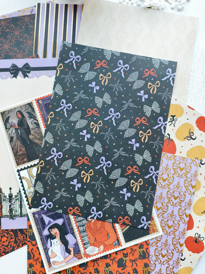 A flat lay of assorted patterned paper sheets and envelopes, with a dark charcoal sheet featuring a repeating motif of purple bows, orange ribbons, and gray leaves centered over other coordinating Halloween-themed papers.