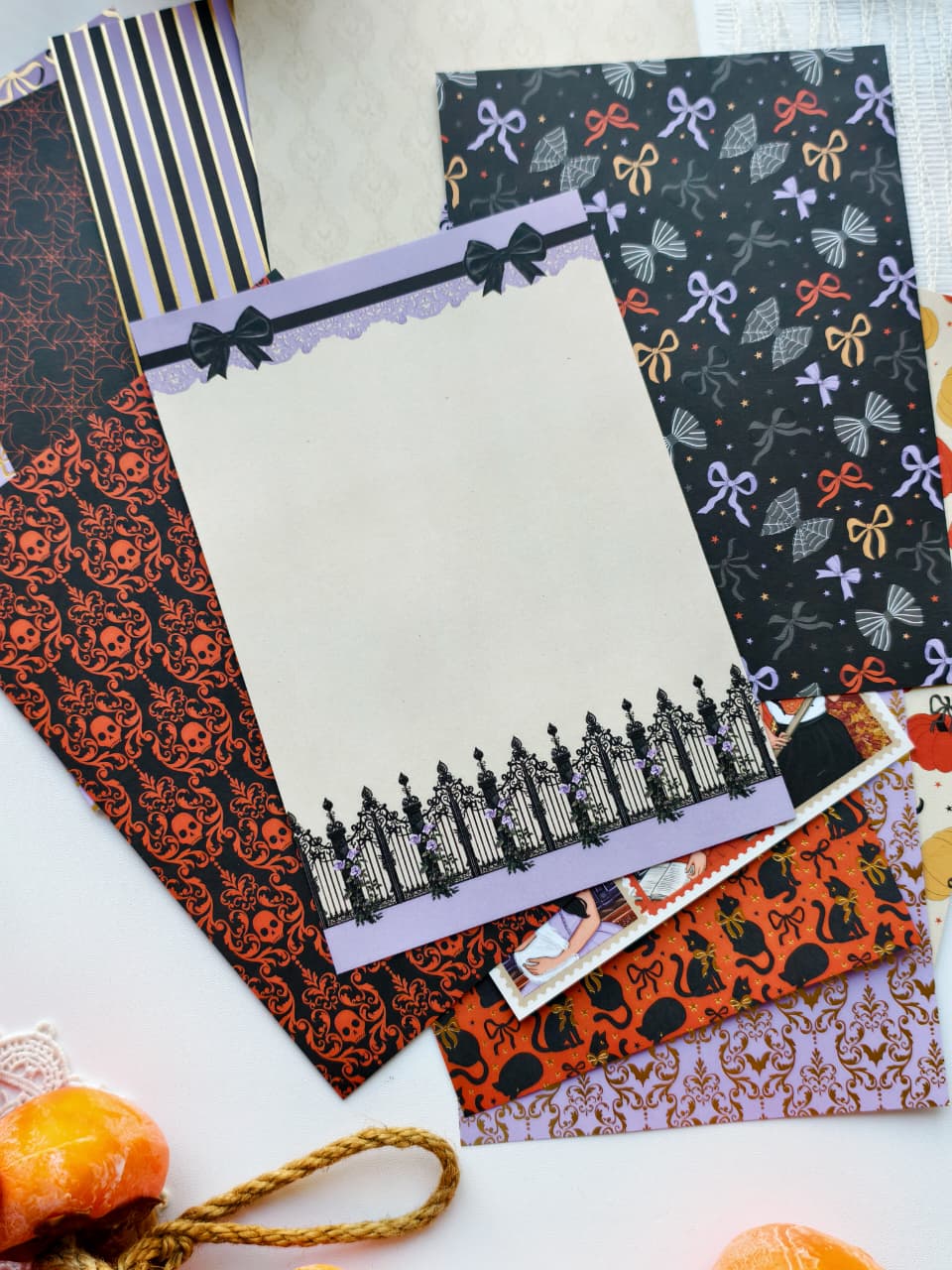 A flat lay of Halloween-themed stationery papers and cardstock patterns, including black and orange spiderwebs, black and purple bows, black silhouettes of houses, and orange pumpkins, arranged on a white surface.