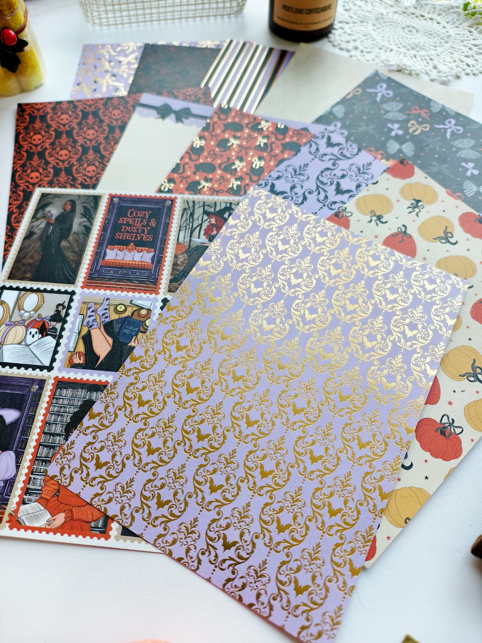 A flat lay of assorted patterned paper sheets on a white surface, including a lavender sheet with gold damask foil, a red-and-black damask sheet, a pumpkin print sheet, and vintage-style illustrated stamps.