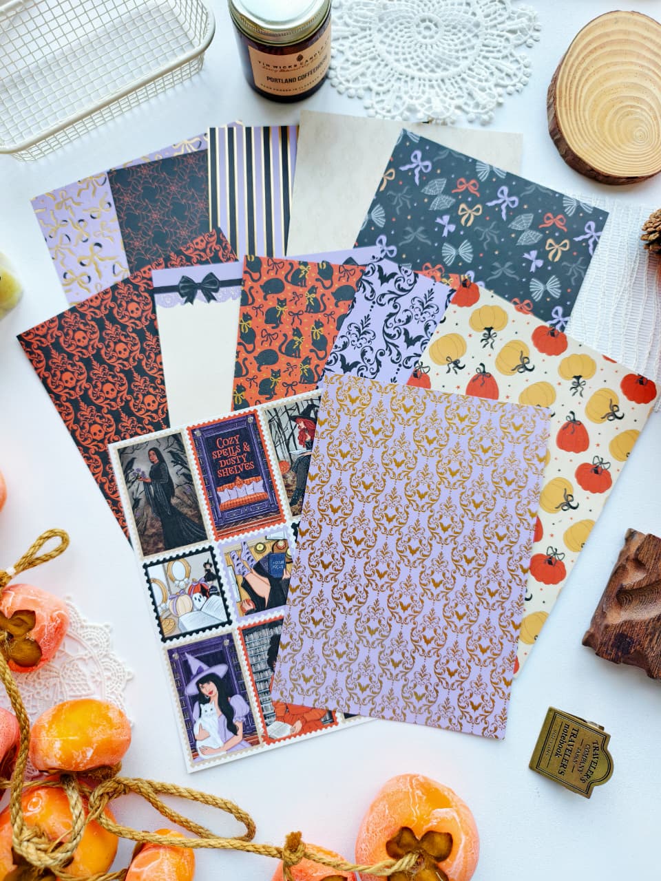 A styled flat-lay of multiple patterned paper sheets and a sheet of illustrated postcards featuring witch and Halloween motifs, arranged on a white surface with autumn props including orange persimmon slices, a lace doily, and a wood slice.