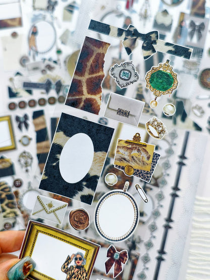 Close-up of a transparent sticker sheet with leopard print pieces, vintage frames, bows, jewelry, and decorative embellishments in neutral and gold tones.