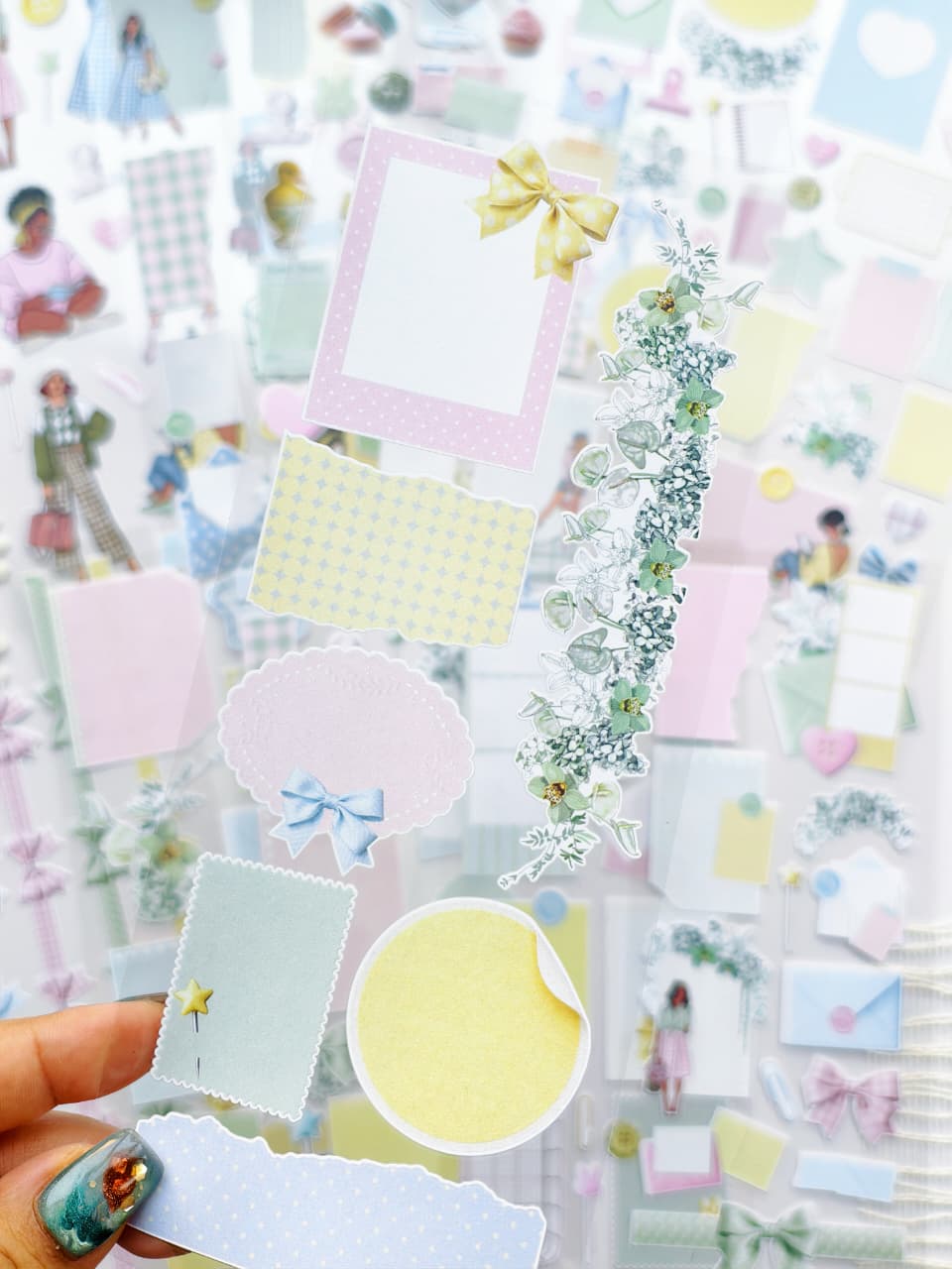 Close-up of pastel transparent stickers with floral garlands, bows, pastel paper scraps, labels, and decorative journaling elements in soft pink, blue, yellow, and green tones.