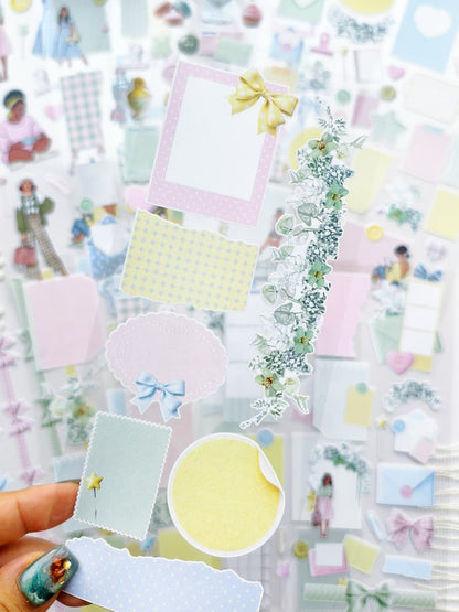 Close-up of pastel transparent stickers with floral garlands, bows, pastel paper scraps, labels, and decorative journaling elements in soft pink, blue, yellow, and green tones.