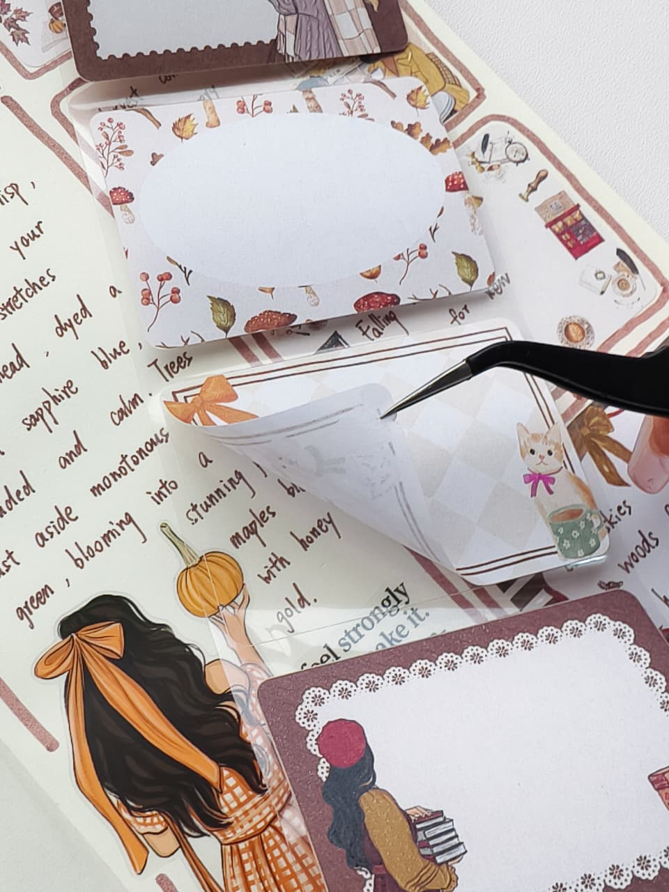 Close-up of tweezers lifting an Autumn Memos Sticky Note from its backing, highlighting the easy peel adhesive and decorative autumn design featuring cozy cats, books, ribbons, pumpkins, and fall foliage.