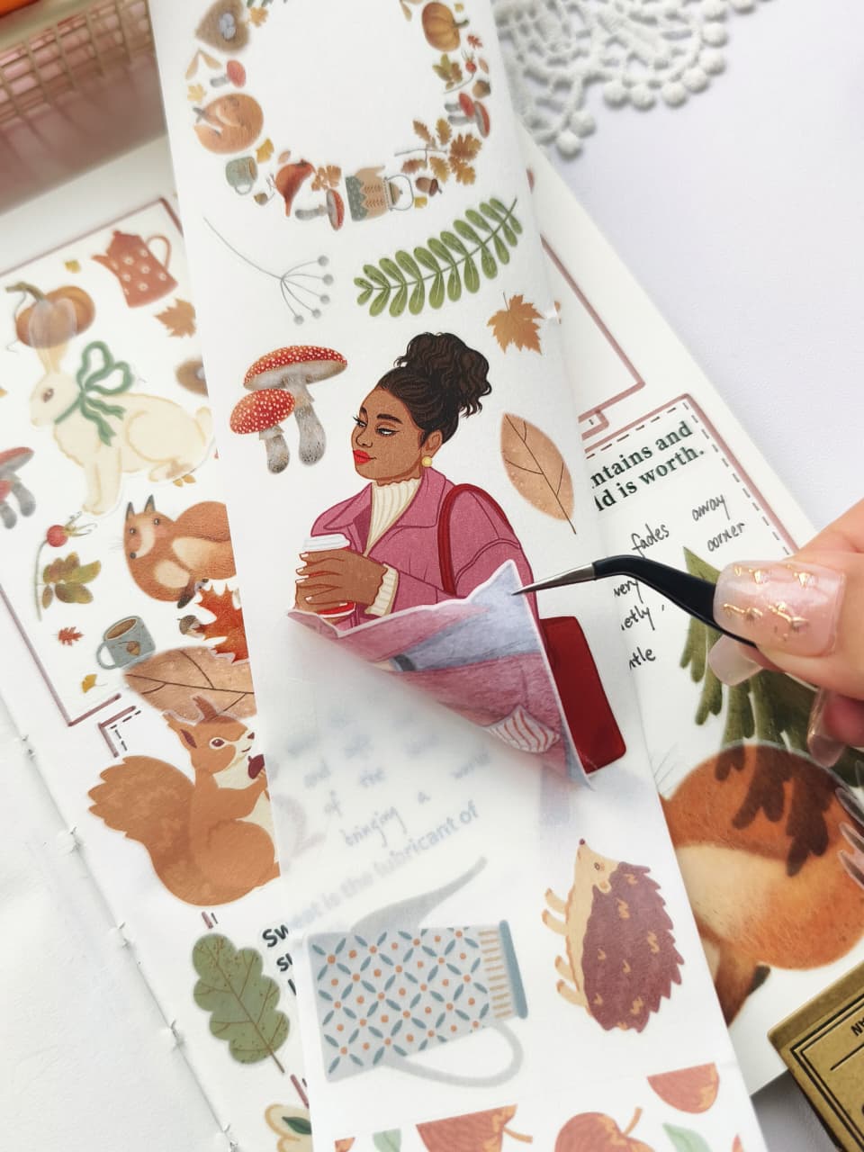 Close-up of tweezers peeling a Foxberry Tales Washi Tape sticker from its backing, highlighting the semi-transparent washi material and easy application of a cozy autumn girl holding a warm drink.