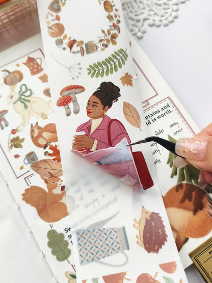 Close-up of tweezers peeling a Foxberry Tales Washi Tape sticker from its backing, highlighting the semi-transparent washi material and easy application of a cozy autumn girl holding a warm drink.