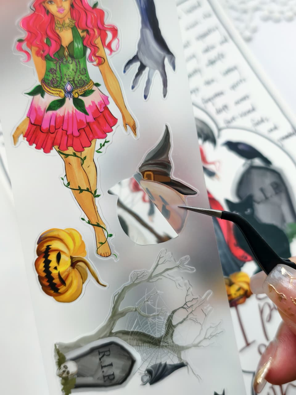 Close-up of tweezers lifting a transparent die-cut Halloween PET sticker from its backing sheet, highlighting the easy-to-peel design with gothic Halloween artwork including a witch hat, pumpkins, gravestone, and bare tree.