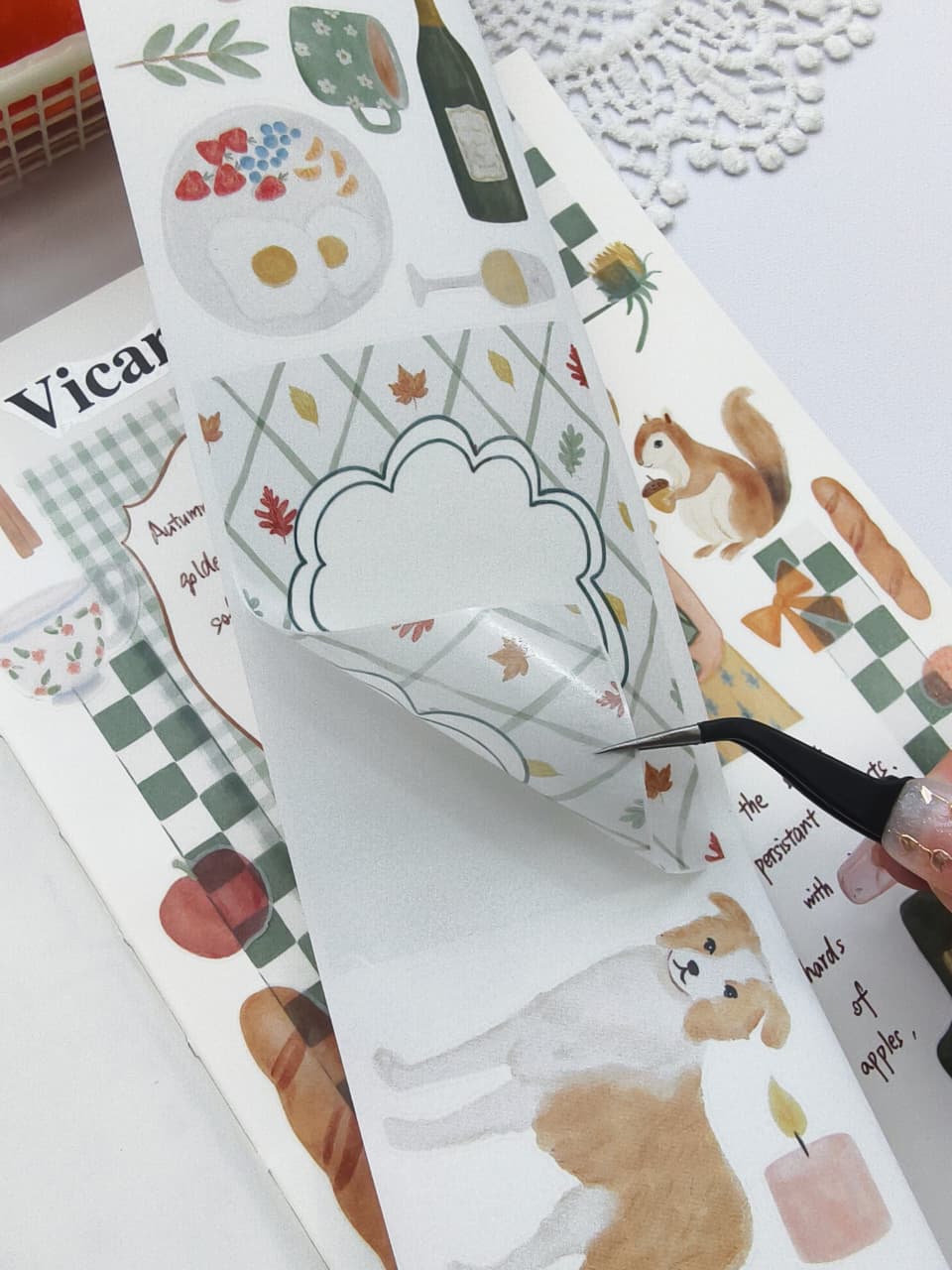 Close-up of tweezers lifting a piece of Cozy Cottage Washi Tape from its backing, highlighting the easy-to-peel design with a decorative autumn journaling label, watercolor leaves, candle, and puppy illustrations.