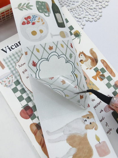 Close-up of tweezers lifting a piece of Cozy Cottage Washi Tape from its backing, highlighting the easy-to-peel design with a decorative autumn journaling label, watercolor leaves, candle, and puppy illustrations.